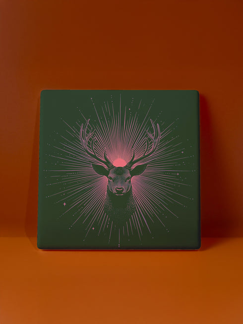 Ceramic coasters in round and square shapes featuring a detailed deer head with majestic antlers against a deep forest green background. A pink-tinged radiant sunburst pattern emanates from behind the deer's head, with delicate stars scattered throughout the design. Cork backing protection.